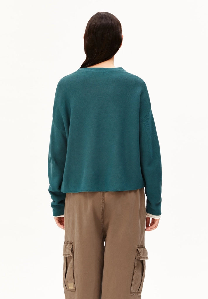 Back view of a woman wearing the ARMEDANGELS Canada Merinaa Links Links Knitted Sweater in Petrol Velvet on a white studio background.