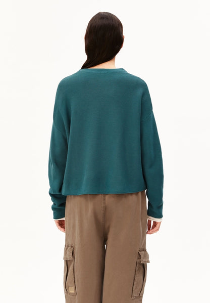 Back view of a woman wearing the ARMEDANGELS Canada Merinaa Links Links Knitted Sweater in Petrol Velvet on a white studio background.
