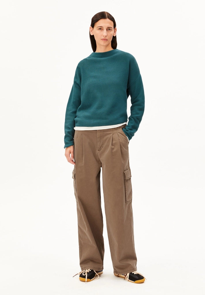 Full-length view of a woman in the ARMEDANGELS Canada Merinaa Links Links Knitted Sweater in Petrol Velvet, with brown cargo trousers and sneakers on a white background.