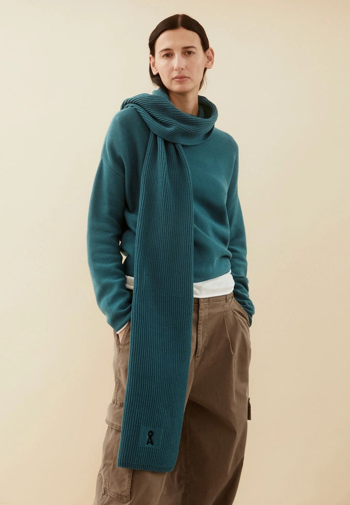 Woman wearing the ARMEDANGELS Canada Merinaa sweater in Petrol Velvet with a matching ribbed scarf, standing against a warm beige backdrop.