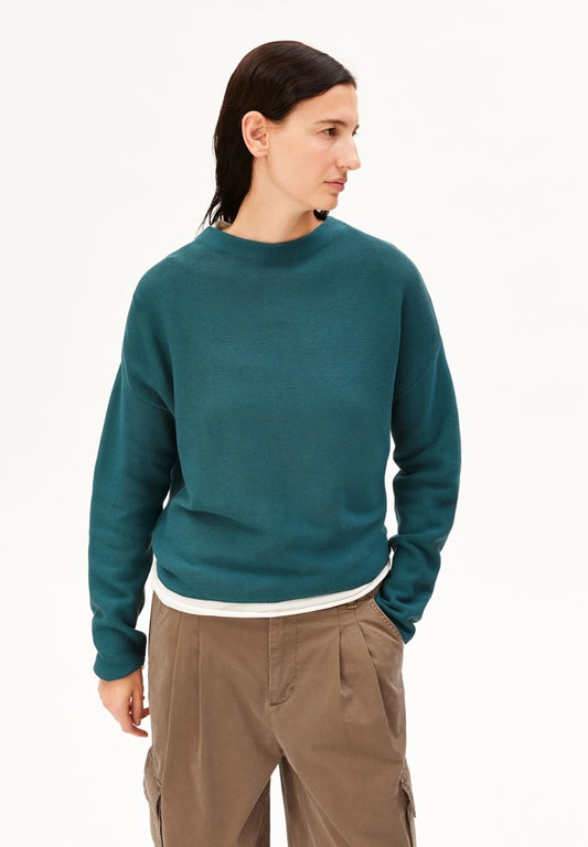 Woman wearing the ARMEDANGELS Canada Merinaa mock neck sweater in Petrol Velvet teal over a white tee, with brown cargo trousers on a white studio background.