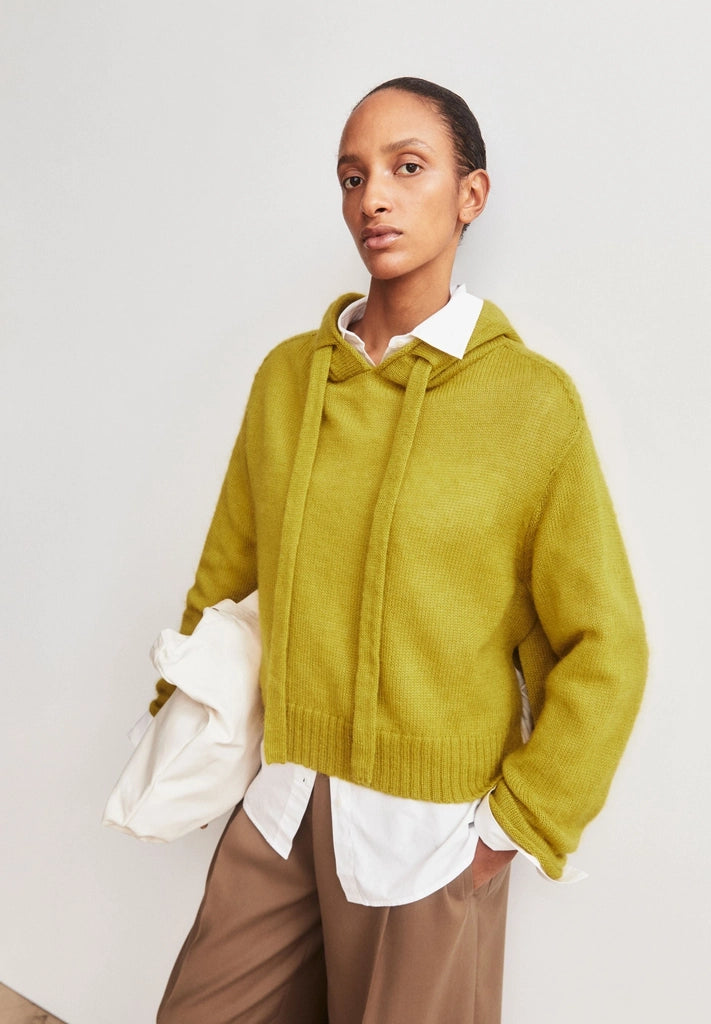 Woman wearing ARMEDANGELS Canada Mohair-Blend Knitted Hoodie in Mosslace with brown trousers, holding a white bag against a light grey wall.