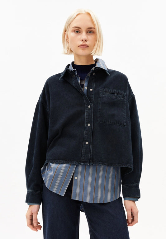 Woman wearing ARMEDANGELS Canada Nalaani Denim Blouse in Rinse over a striped shirt with dark jeans, front view against a white background.