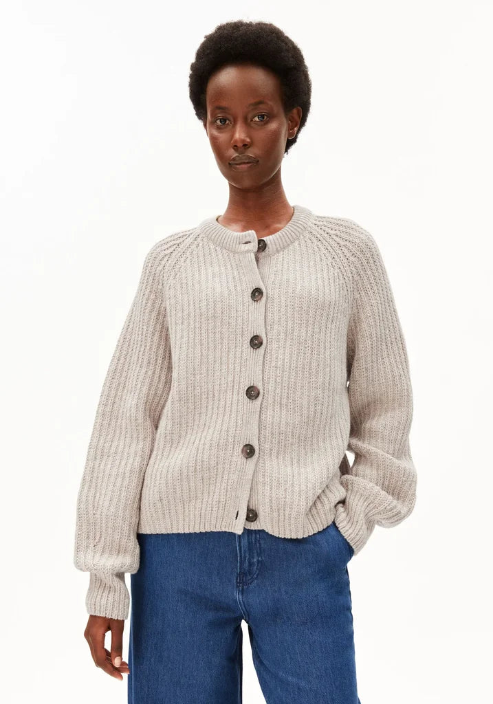 A model wearing a light beige ribbed knit cardigan with dark buttons, paired with blue jeans, against a plain white background.