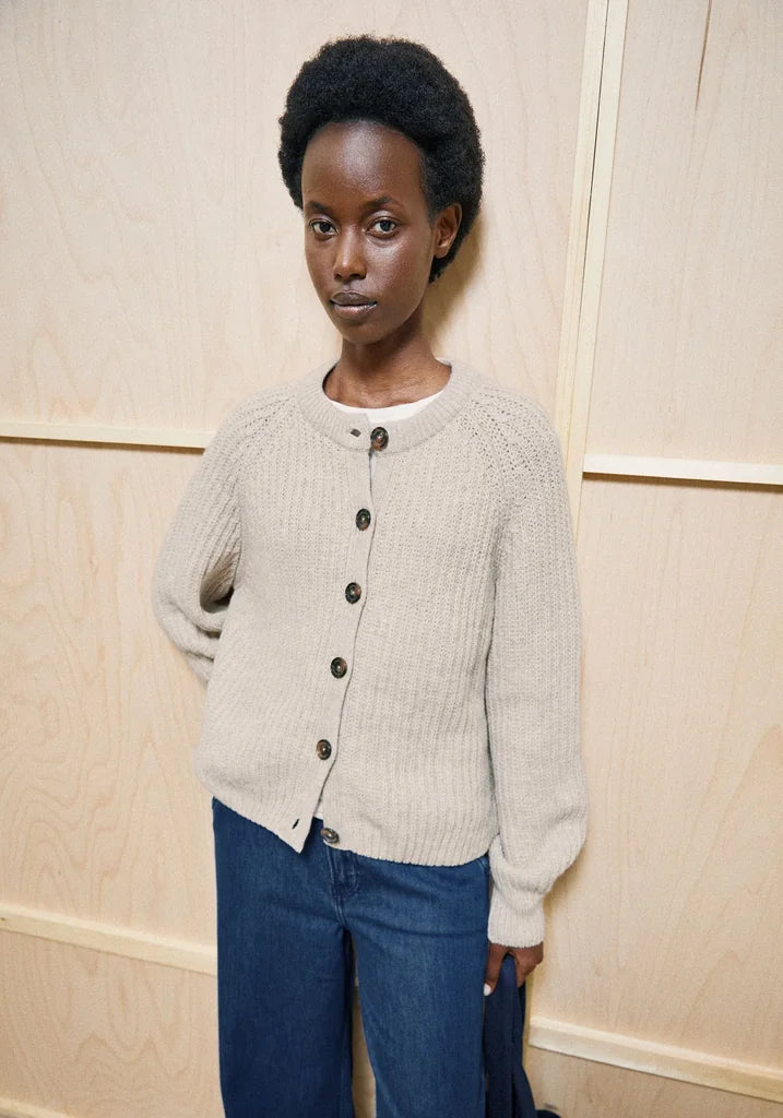 A model wearing a light beige knit button-up cardigan sweater over a white undershirt and blue jeans, standing against a light wood-paneled wall.