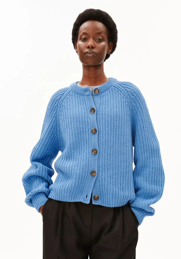 A model wearing a light blue ribbed knit cardigan with brown buttons, paired with black trousers, against a plain white background.