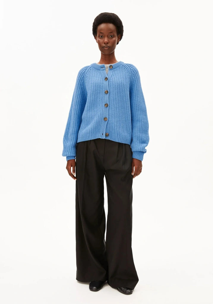 A model stands against a plain white studio background wearing a light blue ribbed knit cardigan with brown buttons and black wide-leg trousers.