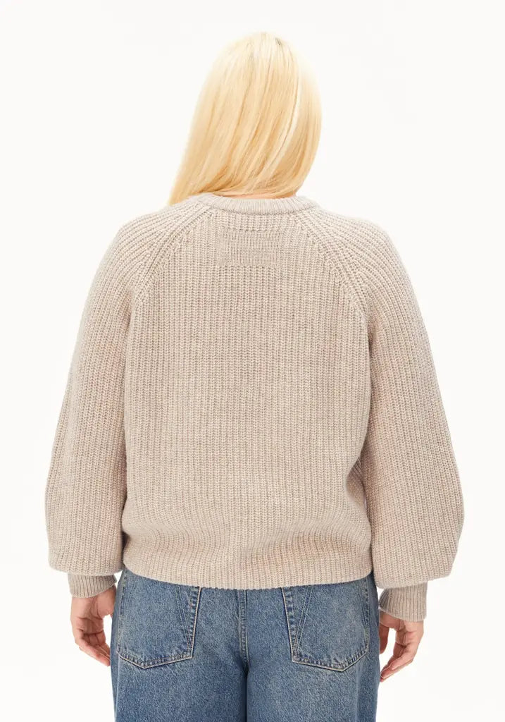 A woman viewed from the back wearing a beige ribbed knit sweater and blue jeans against a plain white background.