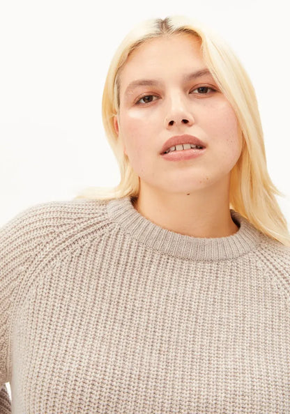 A close-up shot of a woman with blonde hair wearing a beige ribbed knit crewneck sweater against a plain white background.