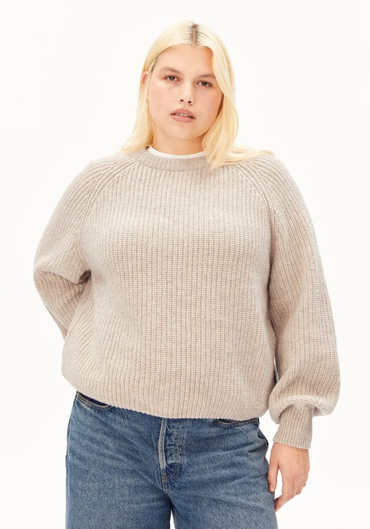 A blonde woman wearing a beige ribbed knit sweater with balloon sleeves and blue jeans, standing against a plain white background.