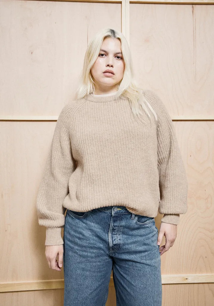 A blonde woman wearing a beige ribbed knit sweater and blue jeans, standing against a light wood-paneled wall.