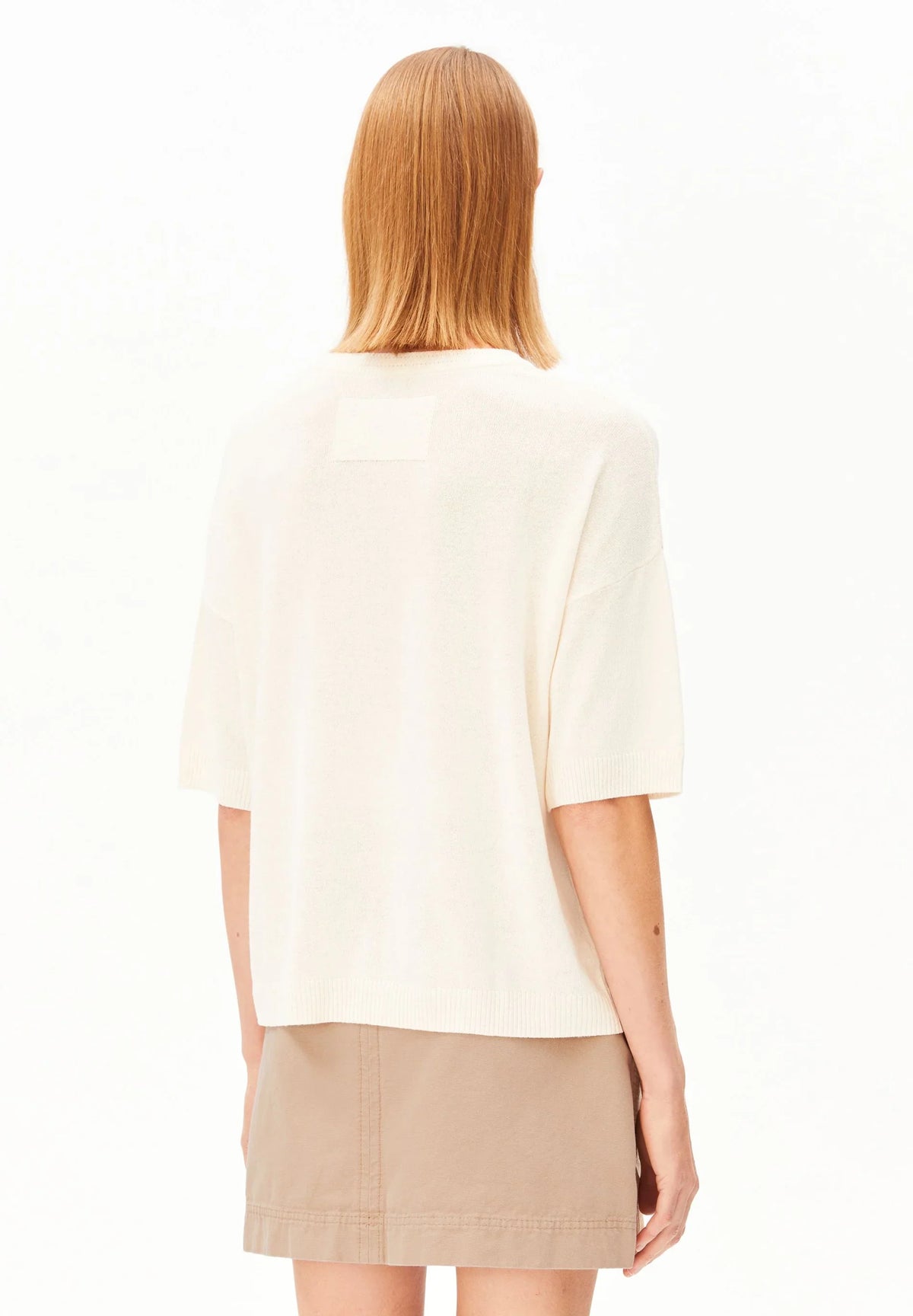 Back view of a woman wearing the ARMEDANGELS Canada Short Sleeve Linen Blend Knit Sweater in undyed cream with a tan mini skirt on a white background.