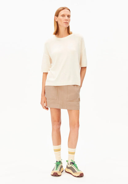 Full-length front view of a woman wearing the ARMEDANGELS Canada Short Sleeve Linen Blend Knit Sweater in undyed cream with a tan mini skirt, striped socks and sneakers on a white background.