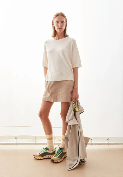 Woman wearing the ARMEDANGELS Canada Short Sleeve Linen Blend Knit Sweater in undyed cream with a tan mini skirt and sneakers, holding a light jacket in a bright white room.