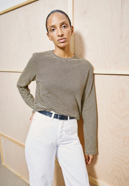 Woman wearing ARMEDANGELS Canada Slub Jersey Striped Longsleeve Shirt in Tinted Navy-Golden Hay with belted white jeans, standing in front of light wood panelling.