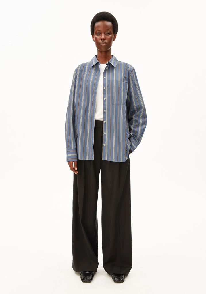 A model stands against a plain white studio background wearing a blue and white vertically striped button-up shirt over a white undershirt, paired with black wide-leg trousers and black shoes.