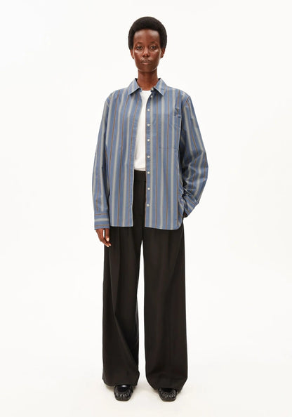A model stands against a plain white studio background wearing a blue and white vertically striped button-up shirt over a white undershirt, paired with black wide-leg trousers and black shoes.