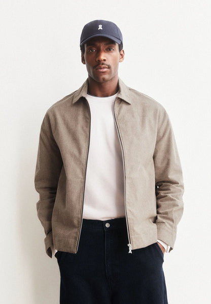 Man wearing ARMEDANGELS Canada Structured Jacket in Honey Reed open over a white tee with a navy cap and dark jeans, against a light grey background.
