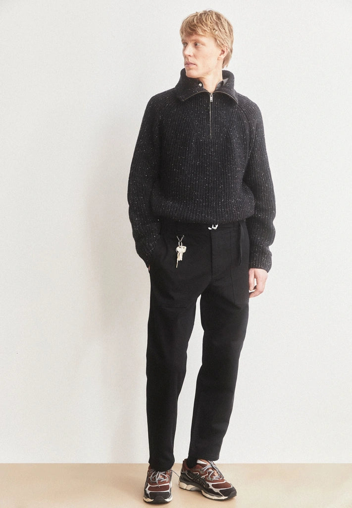 Man wearing ARMEDANGELS Canada Tweed Knit Half-Zip Sweater in black with black trousers, hand in pocket, standing against a light wall.