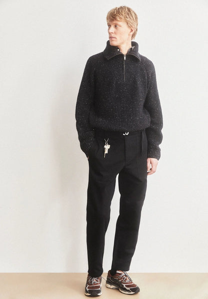 Man wearing ARMEDANGELS Canada Tweed Knit Half-Zip Sweater in black with black trousers, hand in pocket, standing against a light wall.