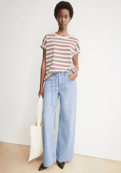 A model stands against a plain off-white wall wearing a short-sleeve crewneck t-shirt with thin red and white horizontal stripes, paired with light-wash wide-leg jeans, black pointed shoes, and holding a white canvas tote bag.
