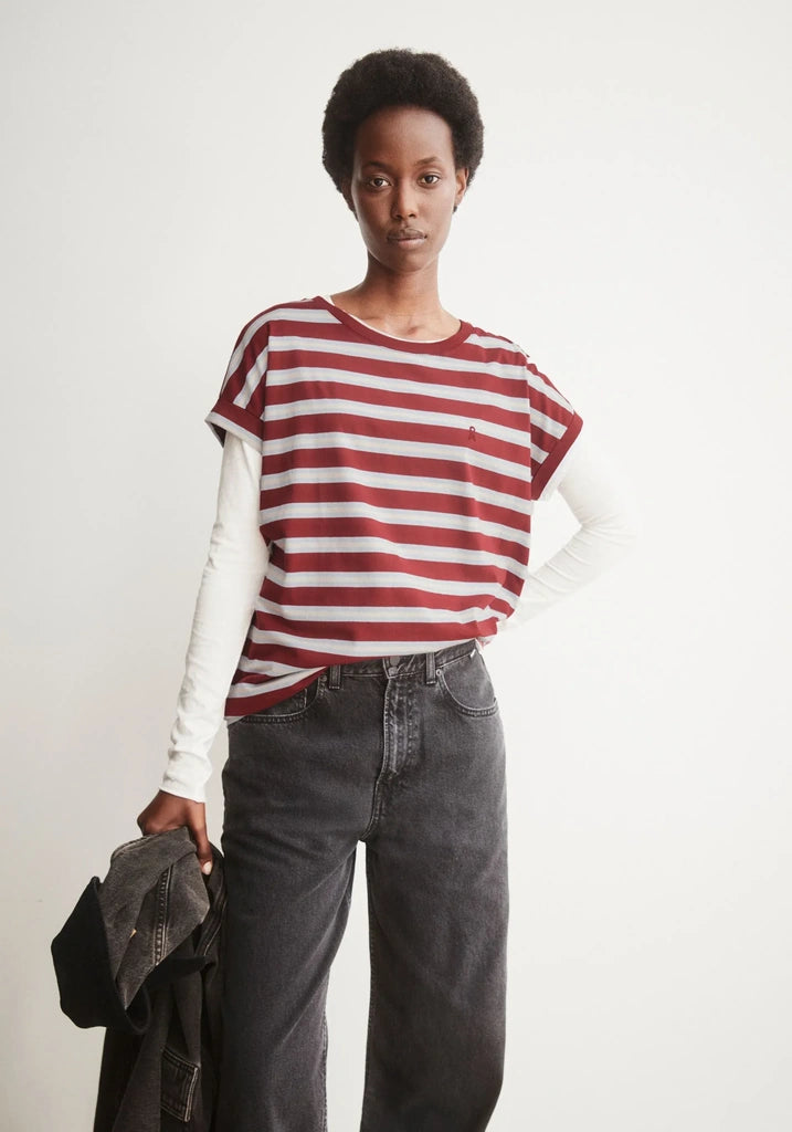A model wearing a red and white horizontally striped short-sleeve t-shirt over a white long-sleeve crewneck shirt, paired with dark gray wide-leg jeans and holding a dark jacket, against a plain white studio background.