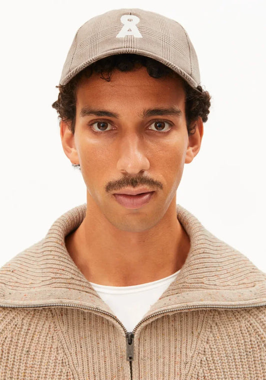 A man wearing a beige plaid baseball cap with a white embroidered logo and a beige ribbed zip-up sweater over a white t-shirt, shot against a plain white background.