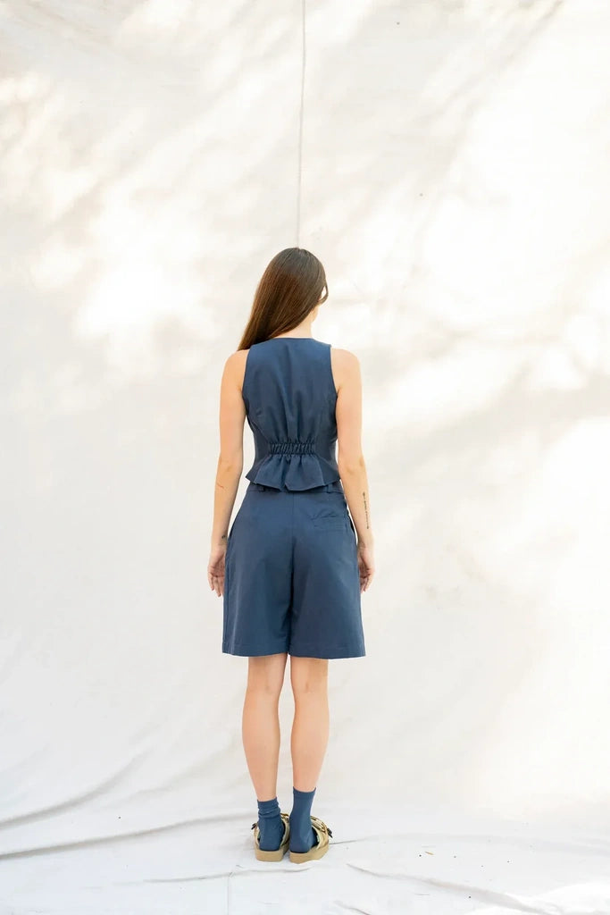 A woman viewed from behind wearing a navy blue sleeveless top with a gathered waist and bermuda shorts, standing against a white draped backdrop with soft leaf shadows.