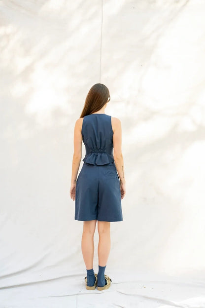 A woman viewed from behind wearing a navy blue sleeveless top with a gathered waist and bermuda shorts, standing against a white draped backdrop with soft leaf shadows.