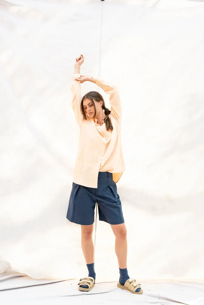 A woman modeling a light peach long-sleeve button-up shirt, navy blue shorts, dark blue socks, and tan slide sandals, posing with her arms raised against a white fabric studio backdrop.