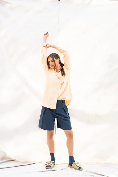 A woman modeling a light peach long-sleeve button-up shirt, navy blue shorts, dark blue socks, and tan slide sandals, posing with her arms raised against a white fabric studio backdrop.