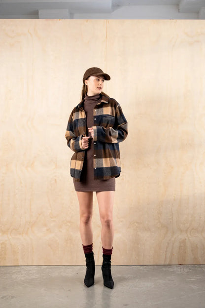 Woman in the BodyBag by Jude Barrington Overshirt in Plaid Quatro and a brown cap, adjusting the cuff, showing the relaxed fit and dropped shoulders over a brown turtleneck dress in a bright studio.