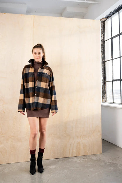 Woman wearing the BodyBag by Jude Barrington Overshirt in Plaid Quatro, a brown, navy and camel plaid wool blend shirt jacket with a point collar, snap front and chest pocket, worn open over a brown turtleneck mini dress with black boots against a plywood studio wall