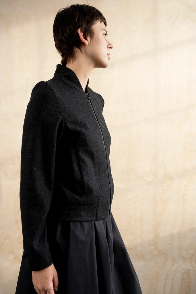 A model in profile wearing a dark charcoal gray zip-up jacket with a high collar and a dark pleated skirt, standing against a textured beige wall.