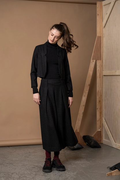 Woman tilting her head in the Bodybag by Jude Nassau Jacket in Charcoal, styled over a black turtleneck with wide-leg trousers in a tan studio.