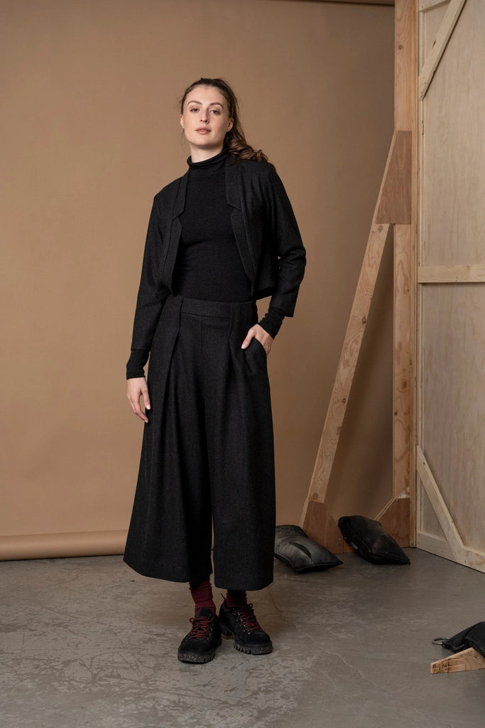 Woman wearing the Bodybag by Jude Nassau Jacket in Charcoal, a cropped wool blend jacket with a sculptural notched open front, over a black turtleneck and wide-leg trousers with red socks and lug-sole shoes in a tan studio.