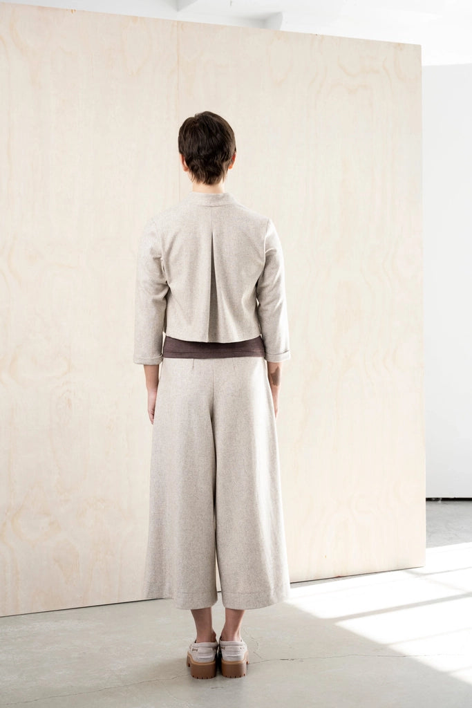 Back view of the Bodybag by Jude Nassau Jacket in Sand, showing the single centre back pleat and the cropped hem above wide-leg trousers.