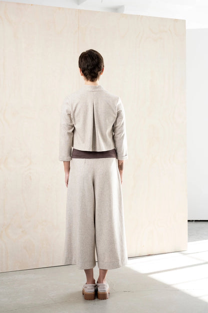 Back view of the Bodybag by Jude Nassau Jacket in Sand, showing the single centre back pleat and the cropped hem above wide-leg trousers.