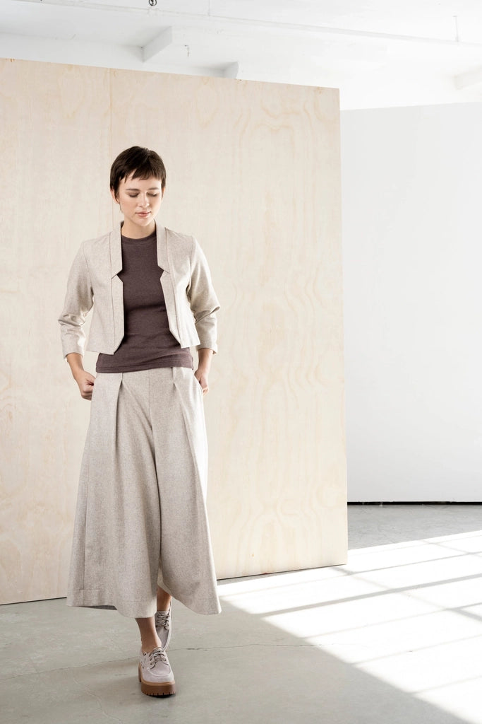 Woman in the Bodybag by Jude Nassau Jacket in Sand with one hand at her hip, showing the cropped, close-fitting shape over high-waisted trousers.
