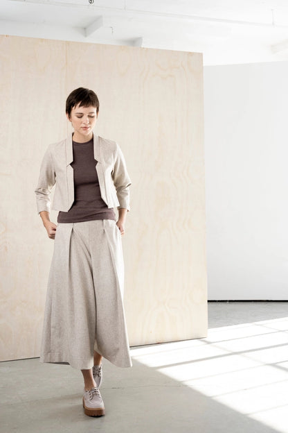 Woman in the Bodybag by Jude Nassau Jacket in Sand with one hand at her hip, showing the cropped, close-fitting shape over high-waisted trousers.