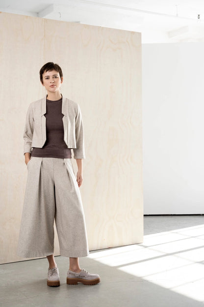 Woman wearing the Bodybag by Jude Nassau Jacket in Sand, a cropped oatmeal wool blend jacket with notched front edges and bracelet-length sleeves, over a brown tee and matching wide-leg trousers in a bright studio with a plywood wall.