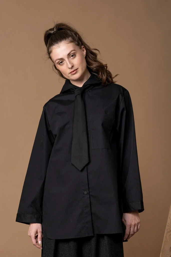 A woman modeling a black long-sleeve button-up shirt with a large bow tie collar, photographed against a solid warm brown studio background.