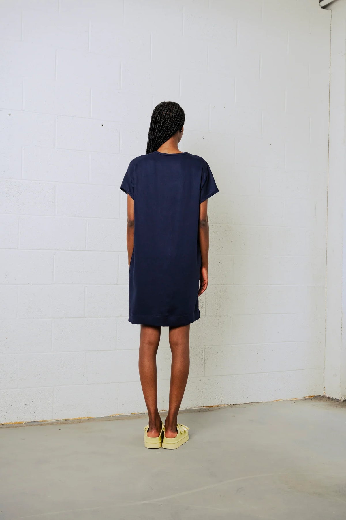 A woman viewed from behind wearing a dark navy blue short-sleeve t-shirt dress, standing against a white brick wall.