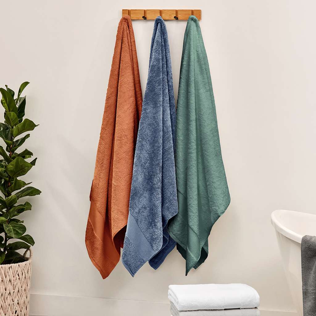 Three textured towels in rust orange, medium blue, and sage green hang from a wooden rack on a bathroom wall, with a folded white towel on the floor and a potted plant to the side.