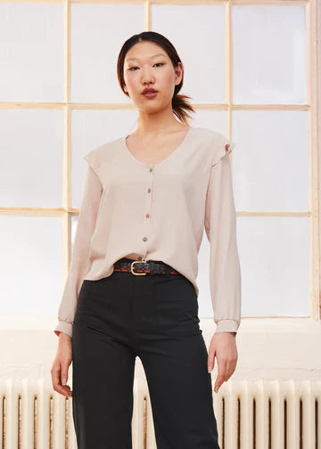 Woman wearing the Dagg and Stacey Aurelia Top in almond with black high-waisted trousers and a brown belt, standing in front of a bright window.