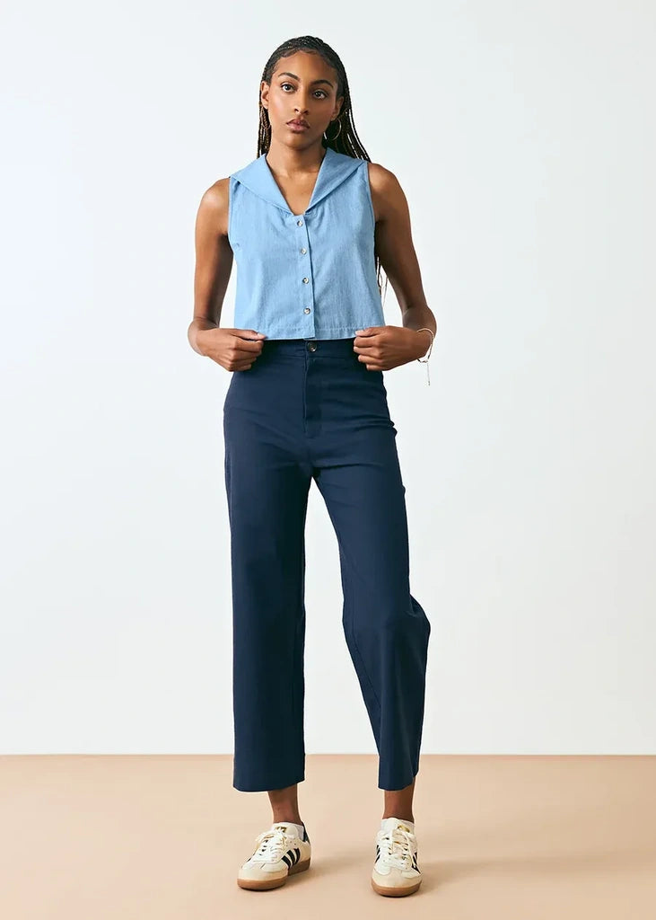 A model stands against a plain light gray studio background wearing a sleeveless light blue button-front top and dark navy cropped pants, with white sneakers.