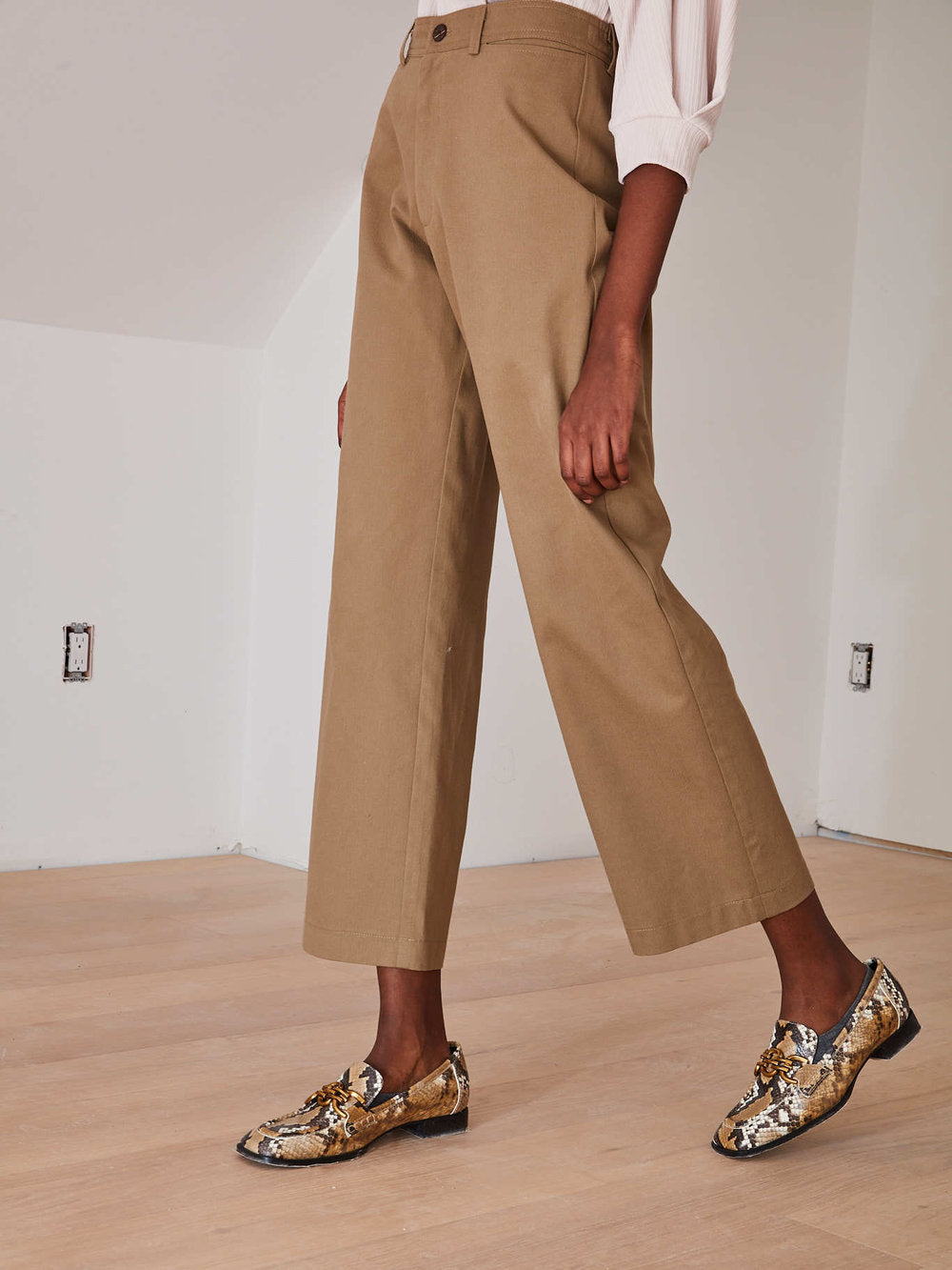 A person wearing tan cropped wide-leg trousers with a light pink top and patterned snakeskin loafers, standing in a minimalist room with white walls and light wood flooring.