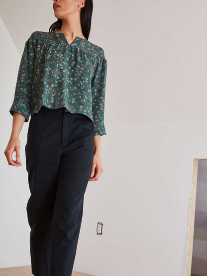 A woman wearing a teal green floral long-sleeve blouse and black narrow-leg trousers, standing in a minimalist white room.