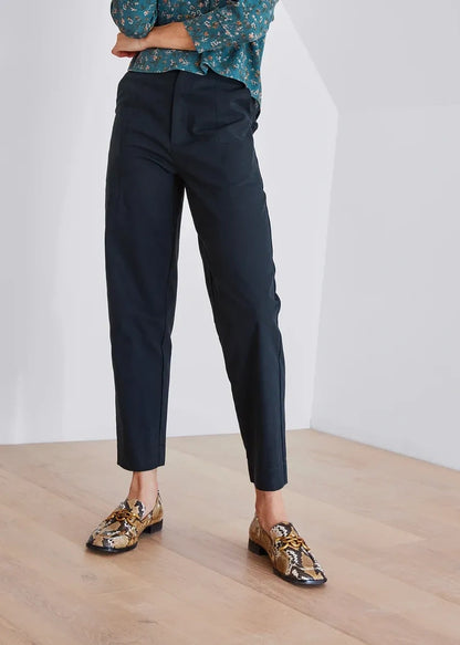 A person wearing slim black trousers, a teal floral blouse, and patterned loafers, standing in a bright indoor room with a light wood floor and white walls.