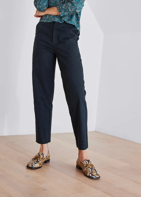 A person wearing slim black trousers, a teal floral blouse, and patterned loafers, standing in a bright indoor room with a light wood floor and white walls.